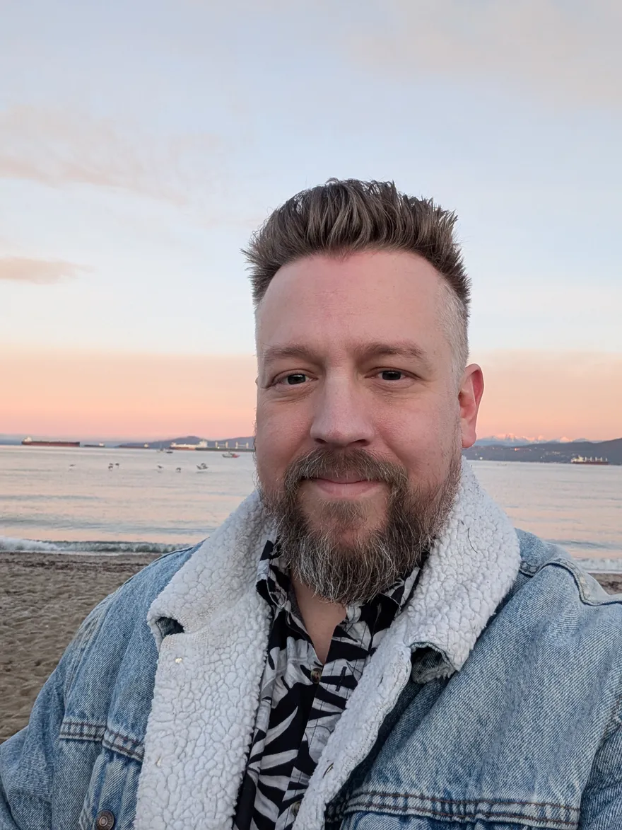 Joel Shinness, a smiling bearded man in a sherpa-lined denim jacket, standing on a beach at sunset with the ocean and distant mountains behind him